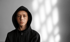 Junger Mann in schwarzem Kapuzenpullover blickt nach vorn, Lichtmuster auf der grauen Wand.