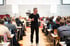 Ein Mann in Schwarz spricht vor einem Publikum mit Tischen in einem großen Saal.