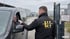 Mitarbeiter von LH Security Service kontrolliert Ausweis eines Autofahrers an einer Werkseinfahrt mit Tor. KI-generiert.