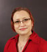 Frau mit rotem Haar, Brille, rotem Hemd und silberner Halskette lächelt.