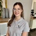 Lächelnde junge Frau mit runder Brille und grauem Poloshirt mit "Fitness für Frauen" Logo.