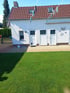 Weißes Einfamilienhaus mit rotem Ziegeldach, drei weißen Türen, gepflasterter Terrasse und einer grünen Rasenfläche.