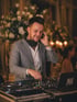 Lächelnder DJ in grauem Anzug mit Kopfhörern am DJ-Pult mit Laptop "PM".