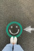 Füße in weißen Turnschuhen vor einem grünen Smiley und einem weißen Pfeil auf Asphalt.