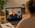Eine Frau sitzt am Schreibtisch vor einem Laptop und führt ein Videotelefonat mit einem älteren, sprechenden Mann.