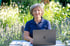 Eine lächelnde Frau mit grauen Haaren sitzt an einem Holztisch mit einem Laptop im Garten.