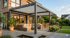 Moderne Pergola mit Lamellendach über einer Terrasse mit Lounge-Möbeln vor einem Haus mit großen Glasfenstern.
