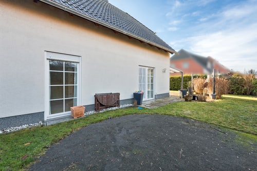 Weiße Hausfassade mit Fenstern und Türen, davor eine asphaltierte Fläche und Rasen, im Hintergrund ein Garten.