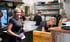 Blonde Frau in schwarzem T-Shirt lächelt und hält eine Flasche in einer belebten Restaurantküche mit Personal.