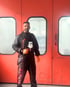 Riccardo Cataldo in schwarzem Overall mit roter Schrift "Andorra" und orangefarbenen Handschuhen hält eine Spritzpistole vor einer roten Tür.
