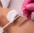 Eine rosa behandschuhte Hand trägt mit einem rosa Applikator Wimpernserum auf die Wimpern einer Frau auf.