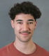 Junger Mann mit dunklen Locken, Schnurrbart und orangefarbenem T-Shirt schaut direkt in die Kamera.