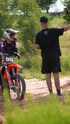 Ein Motocross-Fahrer mit Helm und Schutzkleidung sitzt auf seinem orangefarbenen Motorrad mit der Nummer 98, während ein Mann in Schwarz ihm Anweisungen gibt.