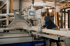 Eine Frau mit Schutzbrille und Gehörschutz bedient eine große, automatische Sägemaschine in einer Werkstatt.