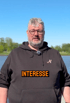 Mann mit Brille und schwarzem Hoodie vor einem See mit eingeblendetem Text „INTERESSE“.