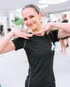 Lächelnde Frau in schwarzem Sport-T-Shirt mit Trainer-Logo posiert mit angewinkelten Armen in einem hellen Boxclub.