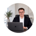 Ein lächelnder Mann im Anzug mit Brille und Kopfhörern sitzt an einem Schreibtisch vor einem Laptop.