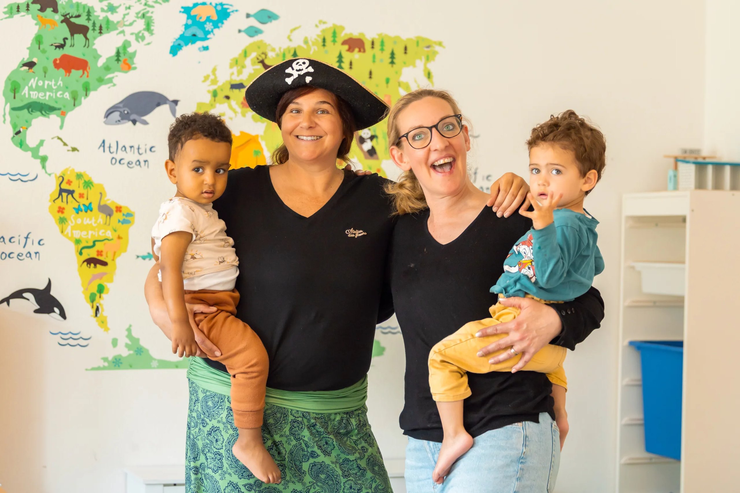 Monia mit Piratenhut und Barbara, beide mit einem Kind auf dem Arm, vor der Weltkarte.