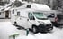 Weißes Wohnmobil parkt im Schnee neben einem kleinen Haus.