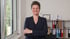 Frau mit kurzen Haaren und dunklem Blazer lächelt, Arme verschränkt, im Büro mit Fenster und Aktenregal.