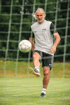 Älterer Mann in Sportkleidung jongliert einen Fußball auf einem Rasenplatz vor einem Tornetz.
