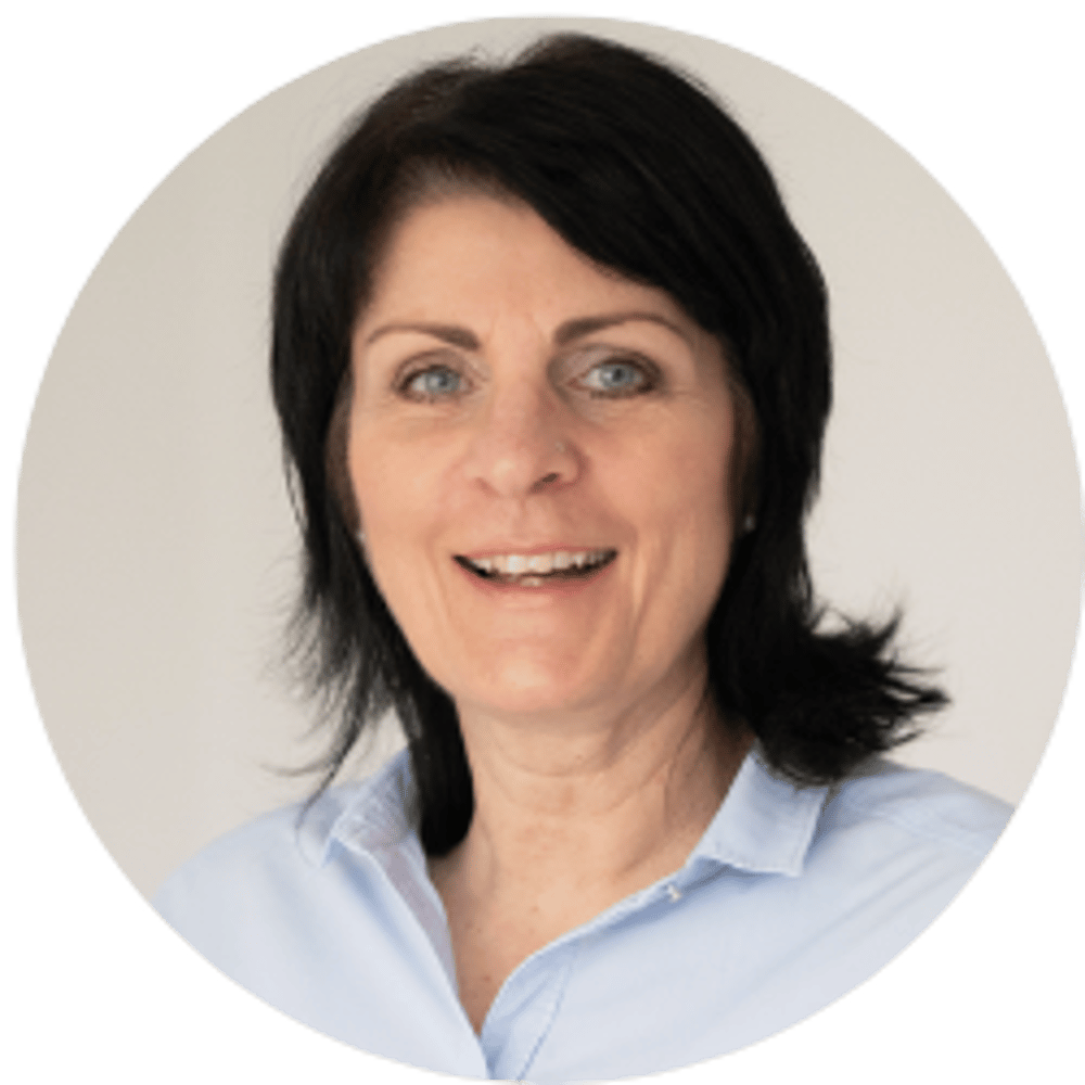 Eine lächelnde Frau mittleren Alters mit dunklen Haaren und blauen Augen trägt ein hellblaues Hemd.