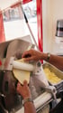 Hände führen Teig durch eine Nudelmaschine, neben einem Behälter mit geriebenem Käse.