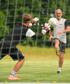 Ein Torwart fängt einen Fußball, der von einem Mann im grauen T-Shirt getreten wird.