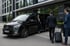 Schwarzer Mercedes-Benz Minivan parkt vor einem modernen Gebäude, während eine Frau aussteigt und zwei Männer mit Gepäck näherkommen.
