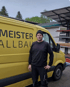 Mann in schwarzer Kleidung und Mütze steht vor einem gelben Transporter mit der Aufschrift "MEISTER ALLBAU".