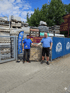 Zwei Männer in blauen T-Shirts stehen vor gestapelten Paletten, einer Steintafel und einem Schild mit Öffnungszeiten.