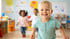 Ein lachender blonder Junge rennt in einem hellen Kindergartenraum auf die Kamera zu.