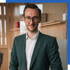 Ein lächelnder junger Mann mit Brille und dunkelgrünem Anzug steht in einem modernen Büro.