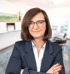 Frau mit braunen Haaren und Brille, trägt einen blauen Blazer und ein weißes Hemd, vor unscharfem Bürohintergrund.