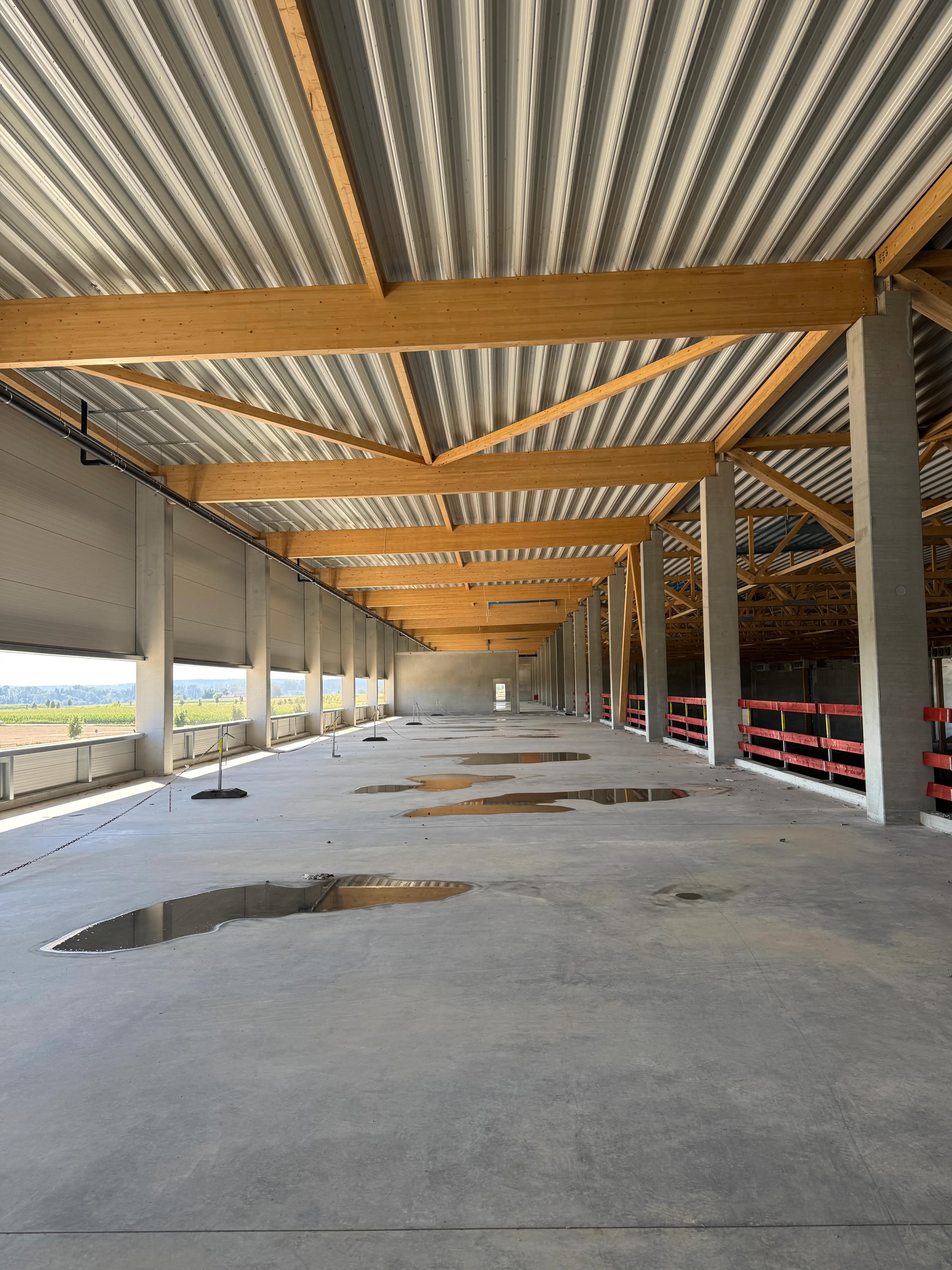 Unfertige Lagerhalle mit Holz- und Betonstützen, gewelltem Metalldach und Pfützen auf dem Betonboden.