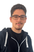 Ein junger Mann mit Brille und Ziegenbart trägt einen schwarzen Kapuzenpullover über einem schwarzen T-Shirt.