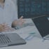 Person am Schreibtisch mit Laptops, Dokumenten und Datenmonitoren gestikuliert während einer Analyse.
