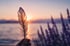 Eine Hand hält eine Feder vor einem See bei Sonnenuntergang und violetten Lavendelblüten im Vordergrund.