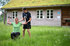 Eine Frau trainiert mit Hanteln im Freien, während ein Mann ihr hilft, vor einem Holzhaus mit begrüntem Dach.