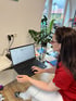 Eine Frau in rotem Shirt und weißer Hose sitzt an einem Laptop auf einem Glastisch und tippt.