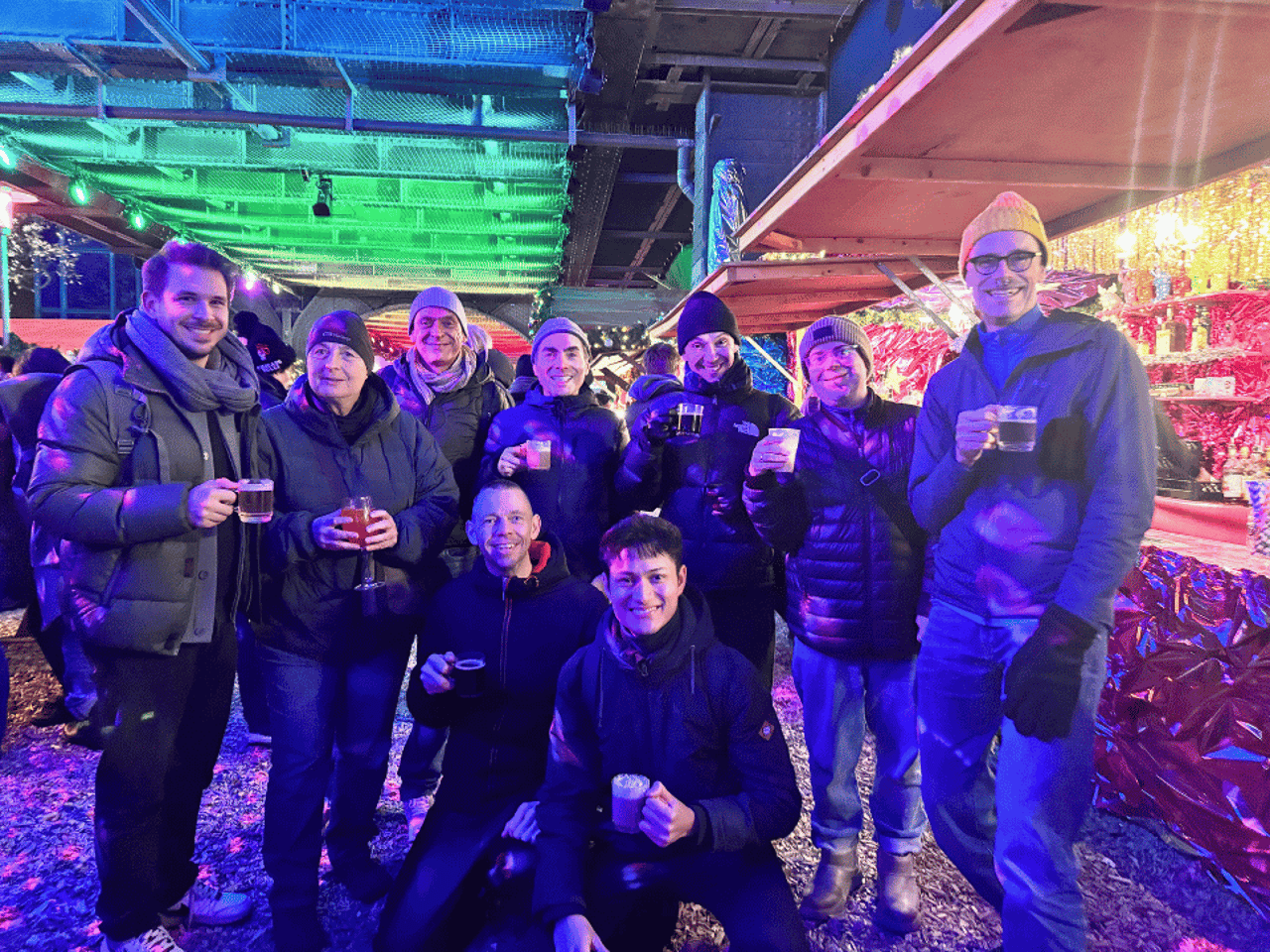 Gruppe von zehn Freunden lächelt und hält Getränke auf einem beleuchteten Weihnachtsmarkt unter einer Brücke.