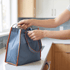 A person’s hands opening a grey insulated lunch bag with orange trim on a white kitchen countertop.
