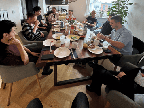 Group of ten people having breakfast at a wooden table in a bright room.