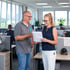 Zwei Kollegen unterhalten sich lächelnd in einem hellen Büro, ein Mann in grauem Polo und eine Frau mit Dokumenten.