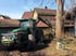 Grüner Traktor mit Kranarm schiebt Äste in einen Häcksler neben einem Baum und Häusern.