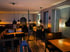 Ein dunkles Restaurant mit Holztischen, schwarzen Stühlen und Pendelleuchten; eine Stehlampe leuchtet rechts.