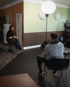 Nia Gralla filmt und interviewt eine weitere Person, die auf einem Stuhl sitzt, in einem Raum mit Licht-Setup.