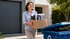 Frau trägt eine Kiste mit Akten zum blauen Recyclingbehälter vor einem Haus mit Garagentor.