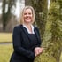 Lächelnde Frau mit blondem Bob-Haarschnitt im dunklen Blazer lehnt an einem moosbedeckten Baumstamm im Freien.