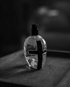 Schwarz-weiße Ansicht einer Parfümflasche mit einem schwarzen Kreuz auf der Vorderseite, auf einer dunklen Oberfläche stehend.
